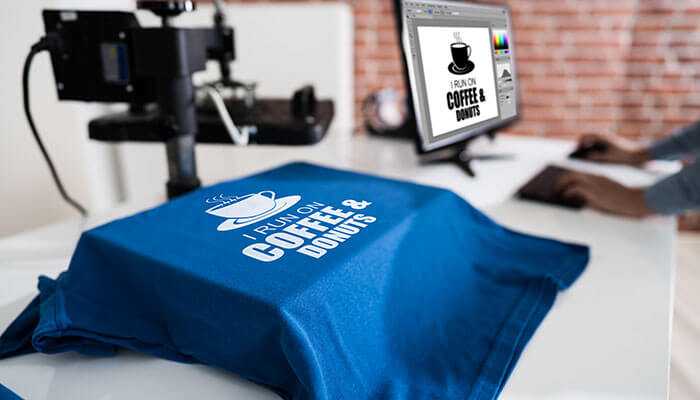 This scene illustrates trends in t-shirt printing, with a blue tee under a heat press as a Fresh Coffee & Donut design appears on the monitor