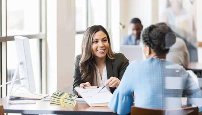 Two women discussing paperwork at a table in a modern office setting, with stacks of cash visible, emphasizing financial planning and loans for gig workers