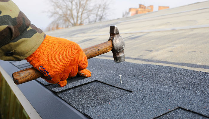 Worker using a hammer to nail shingles on a roof, demonstrating proper techniques to avoid common roofing mistakes during seasonal maintenance.