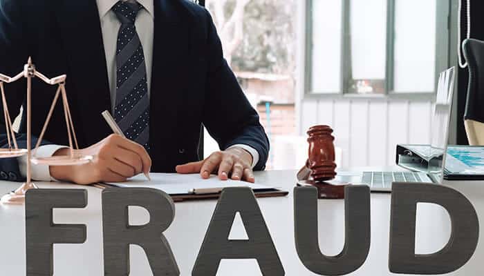 A lawyer in a suit signing documents at a desk with a gavel and the word FRAUD prominently displayed. This image emphasizes the importance of business fraud prevention in legal and corporate settings.