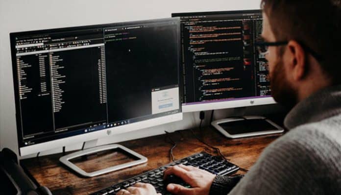 A cybersecurity analyst monitors two screens with code and network logs, illustrating how cybercriminals exploit IT vulnerabilities and system gaps
