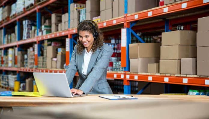 Independent freight agent working on a laptop in a warehouse, managing logistics and freight operations with boxes and inventory in the background.