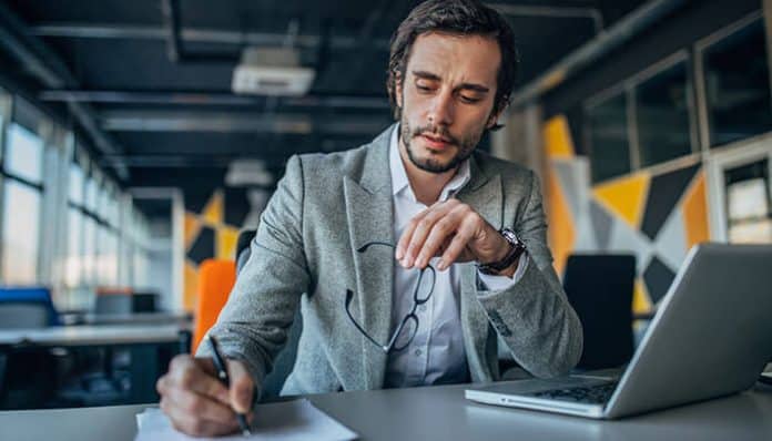 A professional man in a gray suit working at his desk with a laptop and taking notes, representing dedication, professionalism, and how to showcase your expertise effectively in a modern workplace.