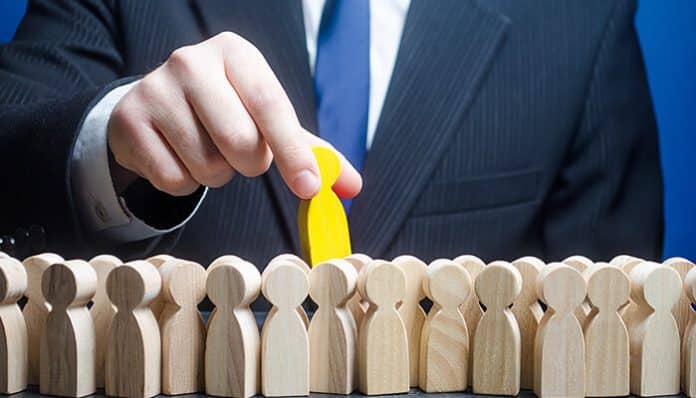 Business professional picking a standout yellow figure from a crowd of wooden icons, representing Best Recruiters for Organizational Growth in hiring the right people.