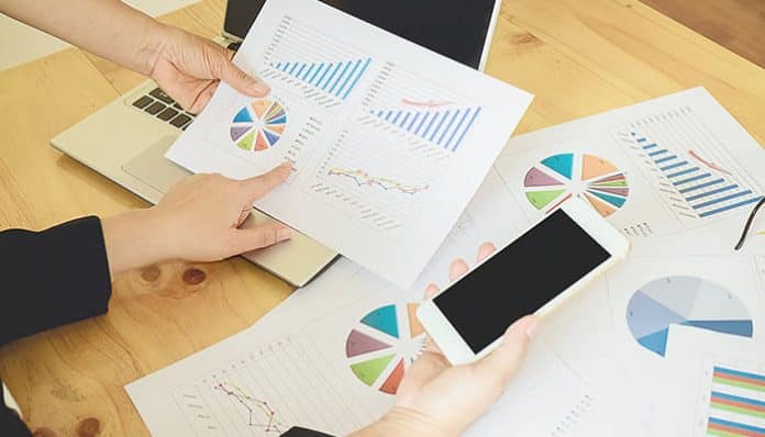 An investor analyzing charts and data on intrinsic value vs market sentiment, using both printed graphs and a mobile device to evaluate stock performance.