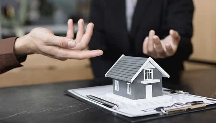 Two professionals discussing property insurance beside a small house model on a clipboard, illustrating key factors that influence commercial building insurance costs.
