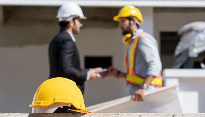 Construction site with workers discussing safety plans while a hard hat rests in the foreground, emphasizing the need for type 2 helmets for better protection