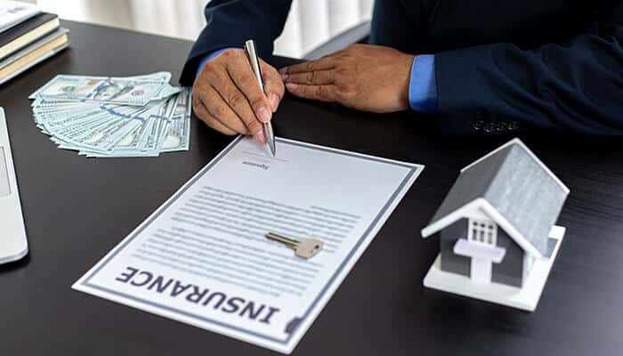 A person signing an insurance document with a house model and cash in the background, emphasizing the role of home investment and protection through insurance.