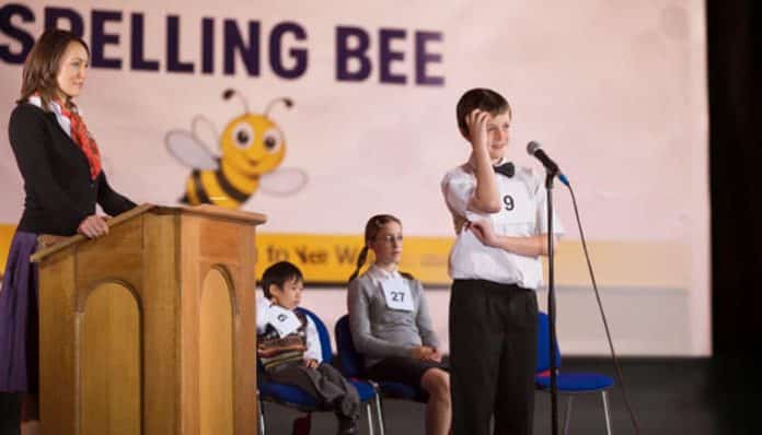 Spelling Bee Buddy concept showing a young student participating in a school spelling bee competition on stage while speaking into a microphone, guided by a judge and audience, representing competitive learning, vocabulary mastery, and structured spelling bee preparation support.
