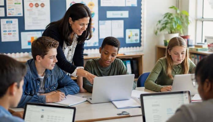 Teacher helps a group of students working on laptops around a classroom table.