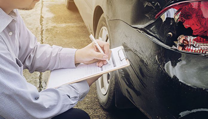 Person writing on a clipboard beside a damaged black car in a parking lot, likely documenting an insurance claim.