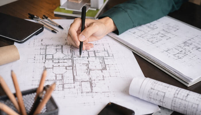 Communication Architecture and Responsiveness - Startupeditor | Startup Editor Architect or designer hand sketching a detailed floor plan on a blueprint, with pencils, rulers, and rolled plans nearby.