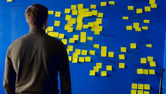 Person with back to camera stands before a blue wall covered in scattered yellow sticky notes during a brainstorming session.