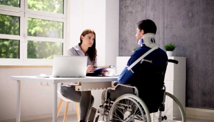 Woman in a gray blouse interviews a man in a wheelchair with a neck brace at a bright office desk with a laptop and documents to discuss.
