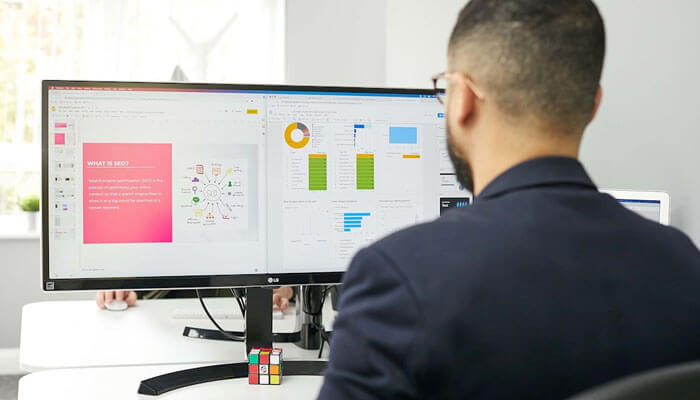 How Marketing Automation Differs from CRM - Startupeditor | Startup Editor Man in a dark suit works at a desk with two monitors showing charts and slides; a Rubik's cube sits on the monitor stand.