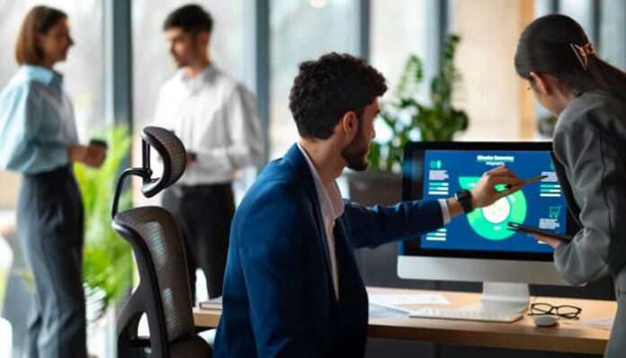 Man in a blue suit points at a monitor displaying charts as a female colleague stands beside him in a modern office. Two people talk in the background.] , wait that has extra bracket.