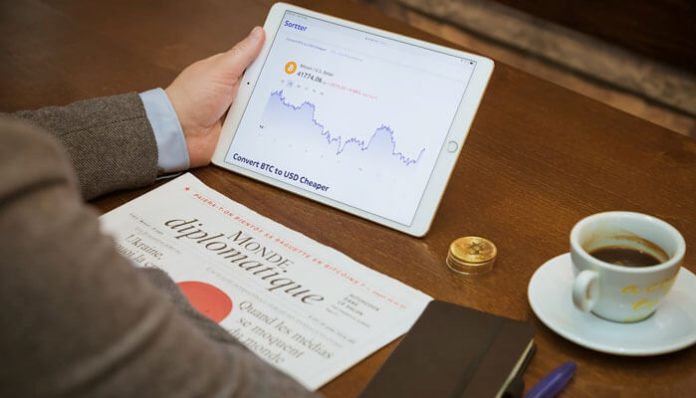Person holding a tablet showing a crypto price chart and downward trend, on a wooden table with a newspaper, a cup of coffee, and coins nearby.