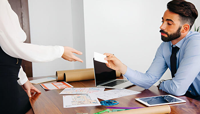 The Risk of Accepting a Settlement Offer Without Legal Advice - Startupeditor | Startup Editor Man in a blue shirt and tie hands a business card to a coworker across a desk, with documents, a tablet, and a laptop on the table.