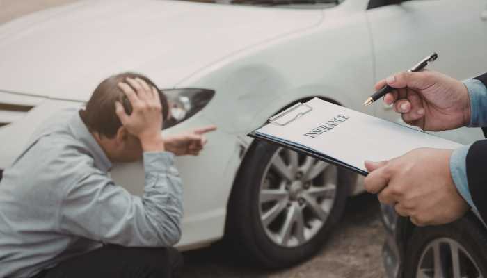 The Serious Impairment Threshold for Michigan Tort Claims | Startup Editor Man sits on the ground beside a damaged white car, holding his head in frustration while an agent fills out an insurance form on a clipboard nearby.
