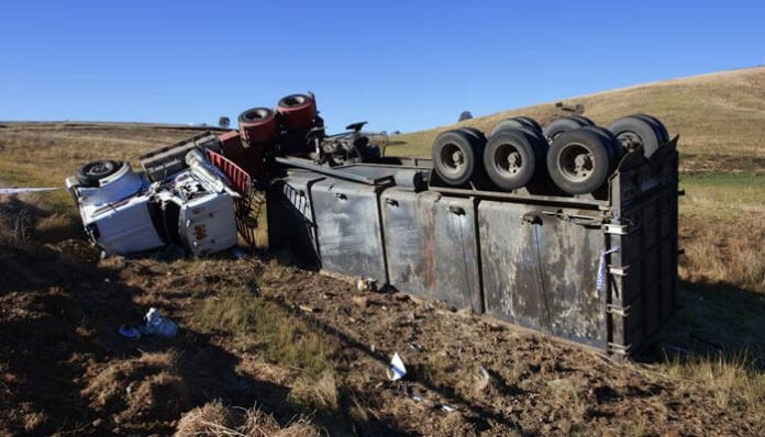 Overturned semi-trailer on its side in a rural field, with wheels up and scattered debris nearby