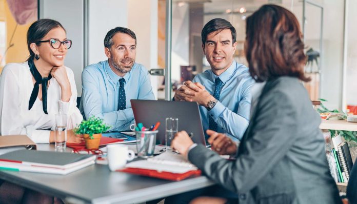 Four business professionals sit around a conference table in an office, looking at a laptop and discussing.