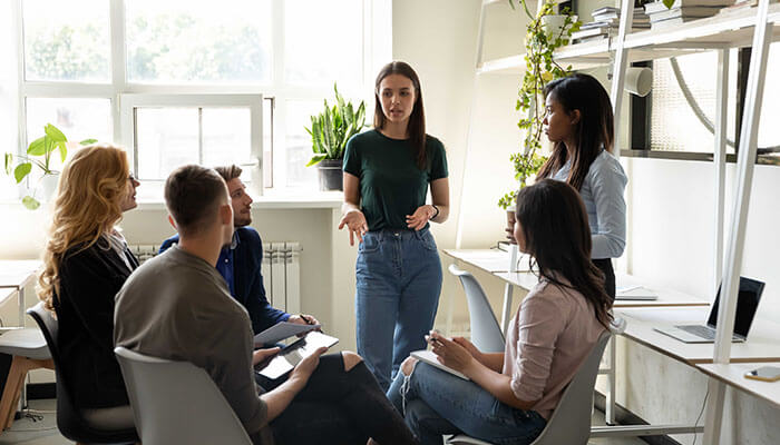 Who Else Gets Caught in the Crossfire - Startupeditor | Startup Editor A woman leads a discussion in a bright office, surrounded by five coworkers seated in a circle.