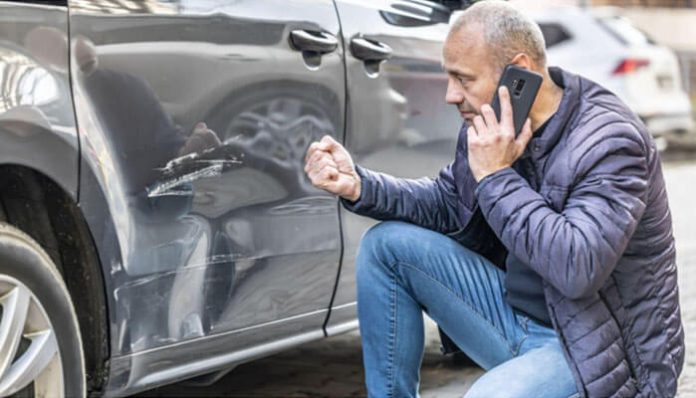 Man crouching beside a dark car, talking on his phone, as he examines a scratched dent on the driver’s door.