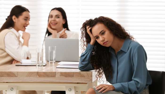 What Anti-Discrimination and Harassment Policies Does a Startup Need | Startup Editor Woman in a blue blouse sits at a desk with her head in her hand, looking stressed, while two coworkers smile in the background near a laptop.
