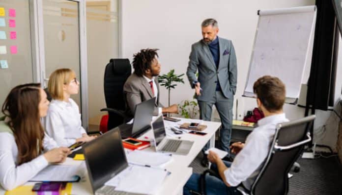 Team meeting in a bright office: five colleagues around a table with laptops as a man in a suit leads a discussion near a flip chart.