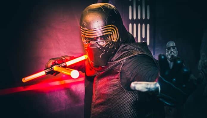 Cosplayer dressed as Kylo Ren wielding a red double‑bladed lightsaber in a dark Star Wars–inspired scene, with a stormtrooper in the background.