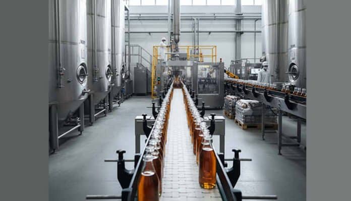 Bottling plant conveyor with amber bottles moving along a stainless steel production line, large tanks on the sides.