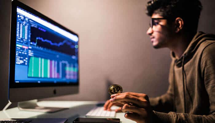 How Might CryptoProNetwork Make Money? - Startup Editor | Startup Editor CryptoProNetwork Adrian concept showing a crypto researcher holding Bitcoin while analyzing cryptocurrency market charts on a desktop computer.