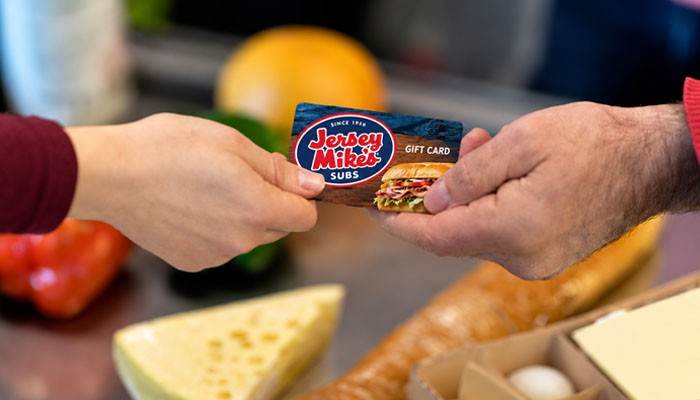 How to Buy a Jersey Mike's Gift Card Online - Startup Editor | Startup Editor Two people exchanging a Jersey Mike's Gift Card over a kitchen counter with fresh sandwich ingredients in the background.